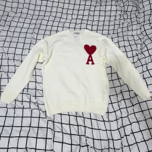 Vit stickad tröja från Ami Paris - Säljer en vit stickad tröja från Ami Paris med röd hjärtlogga och bokstaven A på bröstet. Tröjan har rund hals, ribbade muddar och är långärmad. Perfekt för dig som gillar stilrena plagg med en twist. (Rep)