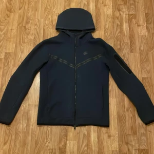 Mörkblå Nike vindjacka med huva - Nike vindjacka i mörkblå med hel dragkedja, huva och diskret logga på bröstet. Jackan har en modern, clean look med svarta detaljer över bröstet och en ficka med dragkedja på ärmen. Tillverkad i mjukt syntetmaterial, perfekt för blåsiga dagar. Den är i storlek S men passar perfekt i M också. Köpt den för 1200kr har använt den några gånger 