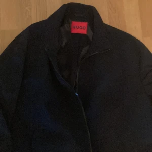 Svart jacka från HUGO med dragkedja - Stilren svart jacka från HUGO med klassisk krage och hel dragkedja framtill. Jackan har släta fickor på sidorna och diskret röd HUGO-detalj vid dragkedjan. Materialet är mjukt och slätt, ger en clean look. Helt ny. Passar M-L, storlek 54 Hugo. Köpte förra hösten för 2990:- men tyvärr för liten nu.