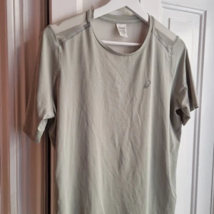 Ljusgrön tränings-tshirt från Asics - Sportig ljusgrå t-shirt från Asics med rund hals och mesh-paneler på axlar och rygg för extra ventilation. Kortärmad och tillverkad i lätt polyester som andas, perfekt för intensiva träningspass. Diskret Asics-logga på bröstet och stilren design. Väldigt mjuk och bra för löpning. 
