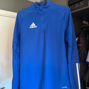 Blå långärmad Adidas half zip tröja - Säljer en blå långärmad tröja från Adidas med vitt logotryck på bröstet och half zip framtill. Tröjan är i funktionsmaterial (AEROREADY) och har en sportig look, perfekt för träning eller fotboll. Snygga detaljer på ärmarna. Funkar för mig fram tills jag blev ca 175 