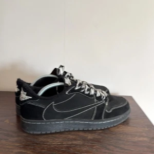 Travis Scott x Air Jordan 1 Retro Low - Säljer ett par svarta Nike Jordan 1 Low med tydliga vita kontrastsömmar och coola svartvita snören. Skorna har en klassisk siluett, rund tå och är gjorda i mocka med detaljer i skinn. Perfekta för dig som vill sticka ut med en unik sneaker.