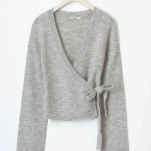 Grå omlottkofta med knyt storlek XS/S - Supermjuk grå omlottkofta med knyt i sidan och v-ringad hals. Koftan är croppad och har långa ärmar, perfekt att slänga på sig över ett linne eller t-shirt. Stilren och enkel att matcha till olika outfits.