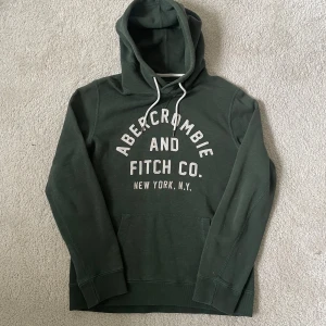 Grön hoodie från Abercrombie & Fitch | XS passar S - Mörkgrön hoodie från Abercrombie & Fitch med vit broderad logga på bröstet och vita snören i huvan. Klassisk känguruficka framtill och ribbade muddar vid ärmslut och nederkant. Perfekt för chill dagar eller när du vill vara extra bekväm.