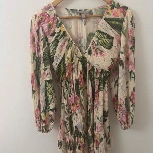 Blommig kortklänning med v-ringning - Superfin kortklänning från H&M med plisserat tyg, v-ringning och trekvartsärmar. Klänningen har ett romantiskt blommönster i rosa, grönt och gult på en ljusbeige bas. Perfekt för sommarens alla tillfällen! Endast använd en gång, så gott som ny