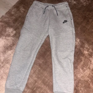 Grå Nike Tech Fleece från Nike, storlek 146-158 - Grå mjukisbyxor från Nike med svart logga på framsidan, snörning i midjan och ribbade muddar vid bensluten. Byxorna har praktiska fickor, varav en med dragkedja. Tillverkade i mjukt material som passar perfekt för chill eller träning.