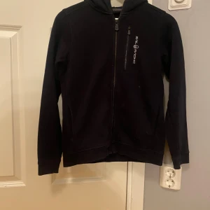 Svart hoodie från Sail Racing med dragkedja - Svart hoodie från Sail Racing med hel dragkedja framtill och en bröstficka med dragkedja. Tröjan har huva och långa ärmar, perfekt för kyliga dagar. Snygg och enkel design med Sail Racing-logga på bröstet.