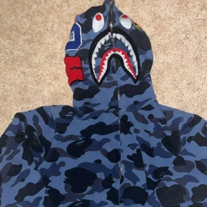 BAPE zip hoodie  - Säljer en ikonisk blå camo hoodie från BAPE med hajmotiv på huvan och dragkedja hela vägen upp. Tröjan har ribbade muddar, coola detaljer och klassisk BAPE-logga på ärmen. Perfekt statement piece för streetwear-stilen.
