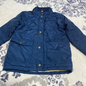 Blå parkajacka från H&M, stl 122 - Mörkblå parkajacka från H&M med flera fickor och snygga guldfärgade knappar. Jackan har hög krage och är helfodrad för extra värme. Perfekt för kyliga dagar och har en klassisk, stilren look.