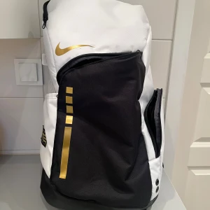 Nike Elite ryggsäck svart/vit/guld - Nike Elite ryggsäck/basketväska i svart och vitt med snygga gulddetaljer och logga. Rymlig modell med flera dragkedjefack, meshficka på sidan och vadderade axelband med transparenta detaljer. saknar dom gummigrejerna på dragkedjorna 