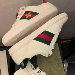 Gucci Ace skor - Gucci Ace sneakers i vitt skinn med klassiska gröna och röda ränder på sidorna och broderad guldfärgad bi. Stl 45, sitter som 44. Aldrig använda bara provade 