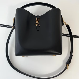 Svart axelväska från Saint Laurent - Snygg svart axelväska från Saint Laurent i slätt skinn med guldiga metalldetaljer och ikoniskt YSL-emblem framtill. Väskan har justerbar axelrem och stilren, boxig form. Perfekt för dig som vill ha en lyxig och tidlös accessoar med exklusiv känsla.