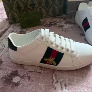Gucci vita sneakers med bi-detalj - Snygga vita sneakers från Gucci i skinn med klassiska gröna och röda ränder på sidan samt broderad guldfärgad bi. Skorna har vit platt sula med Gucci-logga undertill och svart detalj på hälen. Perfekta för dig som vill ha en lyxig och trendig look.