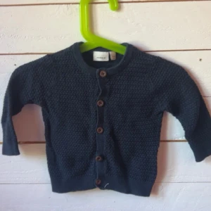 Mörkblå stickad kofta från Name It - Mysig mörkblå stickad kofta från Name It med träknappar framtill. Koftan har långärmad modell och är perfekt för kyligare dagar. Snygg struktur i materialet och klassisk rund halsringning.