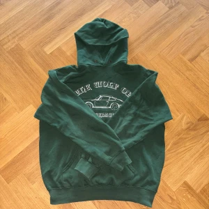 Grön hoodie One of One  - Mörkgrön hoodie från One of One med vit text och en cool bil på bröstet. Klassisk känguruficka framtill, huva med metallöljetter och ribbade muddar. Perfekt för dig som gillar streetwear och vill sticka ut lite extra.