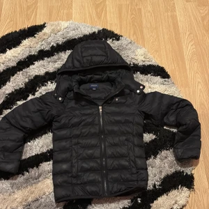 Mörkblå pufferjacka från Gant med huva - Svart pufferjacka från Gant med avtagbar huva och broderad logga på bröstet. Jackan har horisontella sömmar, dragkedja framtill och är perfekt för kyliga dagar. Snygg och enkel design som funkar till det mesta.