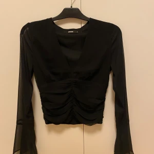 Svart mesh topp från BikBok - Snygg svart topp från BikBok med transparenta meshärmar och djup v-ringning. Toppen har rynkade detaljer framtill som ger en cool look och är croppad i modellen.