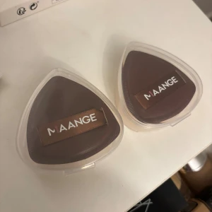 MAANGE makeupsvampar i ask - Två trekantiga makeupsvampar från MAANGE i praktiska genomskinliga askar. Ena sidan är brun med ett band för grepp, andra sidan är persikofärgad. Perfekt för att applicera foundation eller puder jämnt och smidigt.