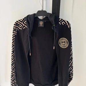 Svart hoodie från Glorious Gangsta M - Svart hoodie från Glorious Gangsta med guldigt emblem på bröstet och feta mönstrade ärmar i svart och beige. Huva med snörning och dragkedja framtill. Grym streetstyle med unika detaljer och bekväm passform.