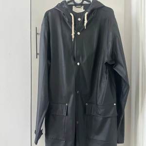 Svart regnjacka från 157 med huva - Svart regnjacka från 157 med huva och vita snören. Jackan har stora fickor framtill, tryckknappar och en stilren look. Perfekt för regniga dagar och enkel att matcha med allt. Materialet är syntet och jackan har en loose passform.