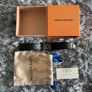 Louis Vuitton svart läderarmband - Snyggt svart armband från Louis Vuitton med ikoniskt LV-spänne i metall och klassiskt monogrammönster på lädret. Armbandet har tryckknapp i silverfärg och kommer med original ask och dustbag. Perfekt accessoar för dig som gillar exklusiv stil.