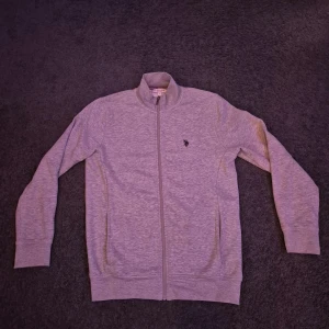 Grå ziptröja från U.S. Polo Assn. - Säljer en grå sweater med fullzip från U.S. Polo Assn Med broderad logga på bröstet. Tröjan har en ståkrage med röd och marinblå randig detalj på insidan samt två fickor framtill. Den har använts en gång och är som sprillans ny! Givetvis äkta! Hör av er vid frågor!