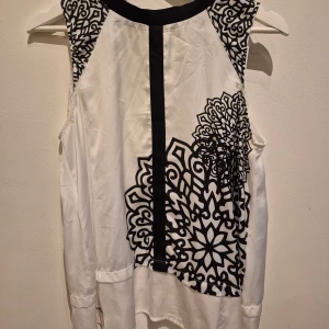 Vit och svart blus från Desigual - Säljer en snygg vit blus från Desigual med svarta grafiska mönster och dekorativ svart kant vid halsen. Blusen är ärmlös och har en guldig dragkedja i nacken. Materialet känns mjukt och lätt, perfekt för en trendig look.