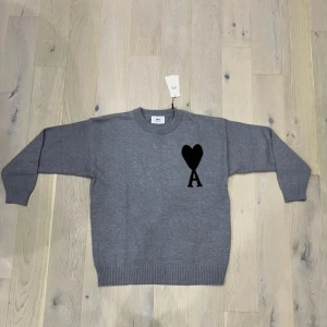 Grå stickad tröja från Ami Paris - Grå stickad tröja från Ami Paris med svart hjärta och bokstaven A broderat på bröstet. Tröjan har rund halsringning, ribbade muddar och är långärmad. Perfekt för dig som gillar stilrena och trendiga plagg.