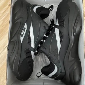 Svarta Dior sneakers med silvriga detaljer - Chunky svarta sneakers från Dior med silvriga detaljer och meshpaneler. Skorna har en kraftig sula, snörning och en modern, sportig vibe. Perfekta för dig som vill sticka ut med exklusiva och trendiga sneakers.