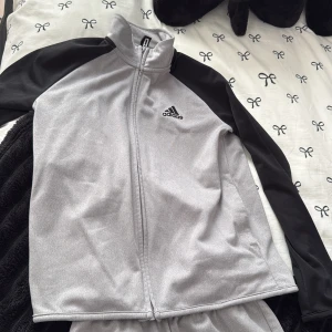 Grå och svart Adidas träningsset - Adidas träningsset med grå zip-jacka och svarta ärmar, samt matchande grå byxor med elastisk midja och Adidas-logga på både jacka och byxa. 