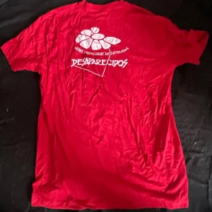 Röd Desaparecidos t-shirt AE Promo XL - Röd t-shirt från American Embroidery Promo i storlek XL med vit tryckt text och blomma på ryggen: 'Desaparecidos' och 'Padres y familiares de detenidos'. Klassisk passform och rund hals, perfekt för dig som gillar statement-plagg.