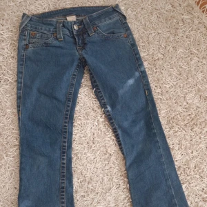 True Religion blå bootcut jeans - Säljer ett par klassiska blå jeans från True Religion med bootcut passform. Jeansen har snygga kontrastsömmar och ikonisk broderad ficka bak. Tillverkade i slitstarkt denim med normal passning och låg midja. Perfekta för dig som gillar en tidlös och cool look.