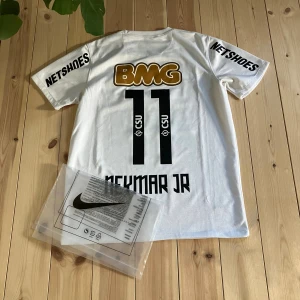 Vit Santos Neymar Jr fotbollströja Nike - Santos FC Neymar Jr matchtröja i vitt med svarta och guldiga tryck. Nummer 11 på ryggen, Neymar Jr-namn, klubbmärke på bröstet och flera sponsorer. Kortärmad modell i lätt och ventilerande material, perfekt för fotboll eller samling.