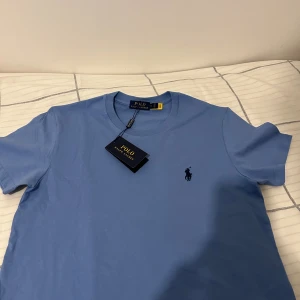Blå t-shirt från Polo Ralph Lauren - Snygg blå t-shirt från Polo Ralph Lauren med klassisk rund hals och broderad logga på bröstet. T-shirten är i mjuk bomull och har en clean, stilren look som passar perfekt till jeans eller shorts.