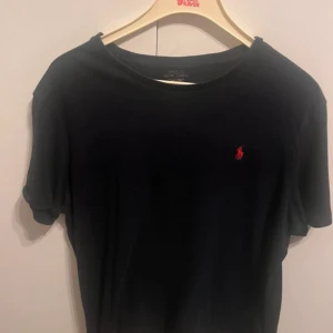 Svart t-shirt från Polo Ralph Lauren - Svart t-shirt från Polo Ralph Lauren med klassisk röd broderad logga på bröstet. T-shirten har rund halsringning och korta ärmar. Tillverkad i mjuk bomull för en skön känsla och enkel stil.