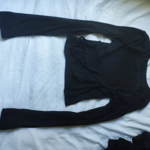 Svart omlottopp med spets från NA-KD - Säljer en svart omlottopp från NA-KD med långa ärmar och snygga knytband i midjan. Toppen är ribbad och har fina spetsdetaljer längs kanterna för extra stilpoäng. Perfekt för dig som gillar trendiga och feminina plagg.