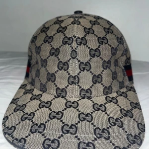 Gucci keps - Snygg Gucci keps i grå med svart GG-monogram över hela kepsen. Klassisk böjd skärm och mörkblå insida. Tillverkad i Italien och gjord av bomullsmaterial. Perfekt för dig som vill ha en lyxig och trendig accessoar. Kom med prisförslag, 