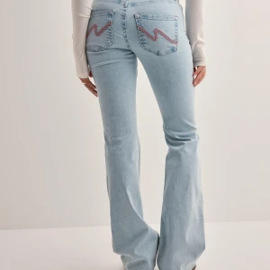 Bootcut low waist ljusblå jeans från Nelly  - Ljusblå bootcut jeans med slitna knän och broderade detaljer på bakfickorna. Jeansen har hög midja och är tillverkade i klassiskt denimtyg. Perfekta för en avslappnad och trendig look.