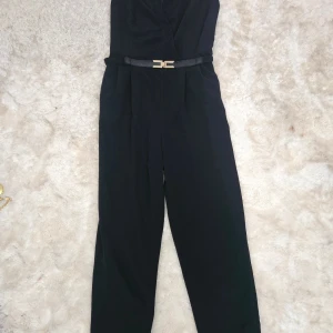 Svart byxdress från River Island - Snygg svart byxdress från River Island, markerad midja och ett smalt bälte med gulddetalj. Byxdressen har dragkedja i ryggen och långa ben, perfekt för en stilren look.