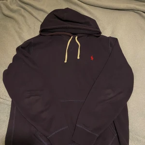 Mörkblå hoodie från Polo Ralph Lauren - Säljer en mörkblå hoodie från Polo Ralph Lauren med klassisk röd broderad logga på bröstet. Tröjan har huva med beige snörning och en stor magficka. Tillverkad i mjuk bomull, perfekt för chill dagar. Passformen är normal. Köpt från boozt.com för 1350 kr och är knappt använd. PRIS ÄR EJ HUGGET I STEN