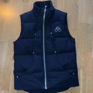Svart väst från Moose Knuckles - Snygg svart dunväst från Moose Knuckles, modell Montreal Vest. Västen har hög krage, två fickor framtill och en tydlig logga på bröstet. Stängs med en grov silverfärgad dragkedja och har en quiltad design som ger en cool look.