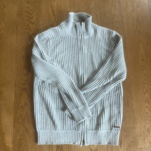 Ljusgrå ribbad kofta med dragkedja från Peak Performance - Säljer en ljusgrå ribbad kofta från Peak Performance med hög krage och hel dragkedja framtill. Koftan är stickad i mjukt material och har långa ärmar samt breda muddar vid ärmslut och nederkant. 