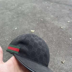 Svart Gucci keps med GG-mönster - Svart keps från Gucci med klassiskt GG-monogram över hela ytan. Kepsen har en grön och röd rand på sidan och justerbar rem baktill. Tillverkad i bomull med detaljer i skinn. Perfekt för dig som vill ha en lyxig och stilren accessoar.