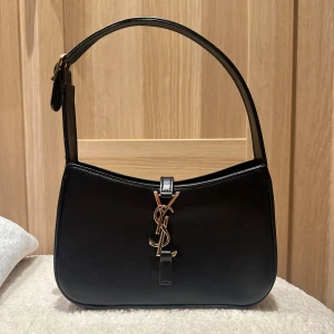 Svart axelväska från Saint Laurent - Snygg svart axelväska från Saint Laurent i slätt skinn med ikoniskt YSL-emblem i guld framtill. Väskan har en stilren, avlång form och justerbart axelband. Perfekt för dig som vill ha en klassisk och trendig accessoar.