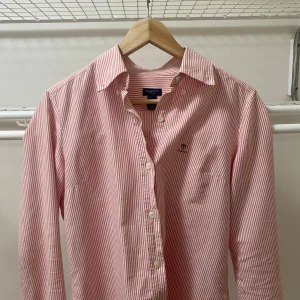 Randig skjorta från Gant i bomull - Klassisk skjorta från Gant med smala röda och vita ränder, broderad logga på bröstet och knäppning framtill. Skjortan har lång ärm, krage och är tillverkad i mjuk bomull. Perfekt för en clean och stilren look.