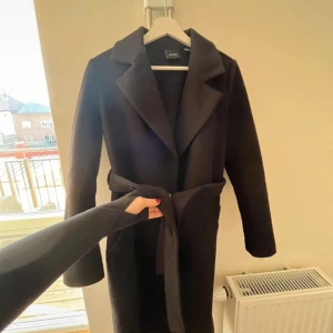 Svart kappa med bälte - Stilren svart kappa från Veromoda med klassisk krage och knytskärp i midjan. Lång modell med raka ärmar och snyggt fall. Perfekt för dig som vill ha en clean och elegant look under höst och vinter. 😇