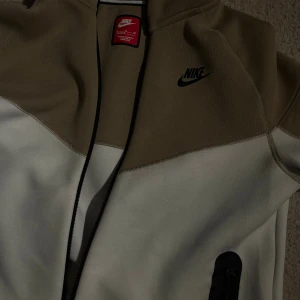 Beige och vit Nike hoodie XL - Snygg hoodie från Nike i beige och vitt med svart Nike-logga på bröstet. Tröjan har huva med dragsko och hel dragkedja framtill. Perfekt för dig som gillar sportig och avslappnad stil. Xl barn passar som xs men