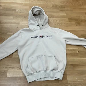 Vit hoodie från Tommy Hilfiger - Vit hoodie från Tommy Hilfiger med broderad logga och text 'EST. 1985' på bröstet. Klassisk känguruficka framtill, ribbade muddar och huva med snörningshål. Perfekt för en chill och stilren look.