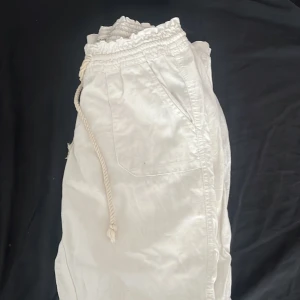 Vita linnebyxor från Roxy med snörning - Säljer ett par vita linnebyxor från Roxy med bred resår i midjan och grov snörning framtill. Byxorna har stora fickor på sidorna och är tillverkade i ett lätt och luftigt linnematerial, perfekta för varma dagar. Ena snöret har blivit lite konstigt där av priset förutom de är skicket väldigt bra då de knappt har använts.