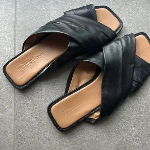 Svarta sandaler från Flattered i skinn - Stilrena svarta sandaler från Flattered med breda korslagda remmar i mjukt skinn. De har fyrkantig tå och platt sula i beige och svart, vilket ger en modern och trendig look. Perfekta för varma dagar när du vill ha både komfort och stil.